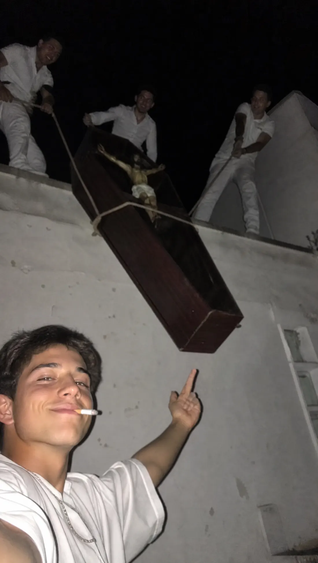 Un chico fuma mientras otros cargan un ataúd por un tejado de noche.